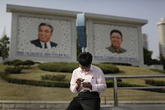 Ein Nordkoreaner sitzt in Pjngjang vor den Gemlden der verstorbenen nordkoreanischen Diktatoren Kim Il Sung (l) und Kim Jong-il (r) und nutzt sein Smartphone.