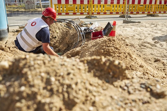 ein Bauarbeiter mit rotem Bauhelm und einer weien Weste mit Vodafone-Logo arbeitet in einem Erdloch und verlegt dort schwarze Kabel