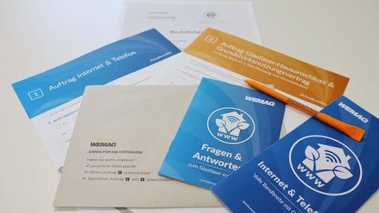 das Bild zeigt verschieden in blau und orange gehaltene Flyer mit weier Aufschrift sowie einen grauen Umschlag der mit WEMAG adressiert ist