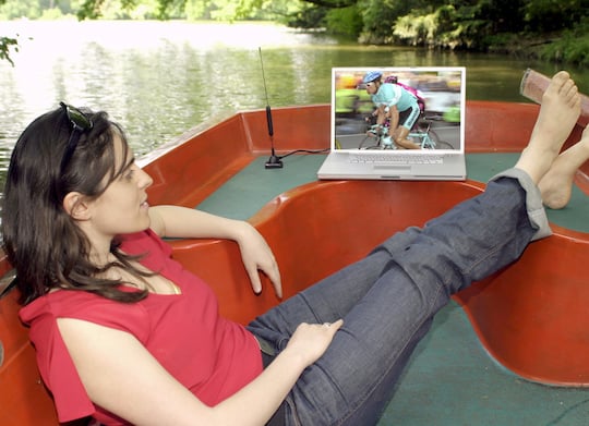 Eine Frau in dunkler Jeans und rotem Oberteil liegt an einem See auf einer Bank und blickt auf einen Laptop, auf dessen linker Seite eine kleine Stabantenne steht. Auf dem Display des Laptops luft ein TV-Programm, das gerade ein Radrennen bertrgt.